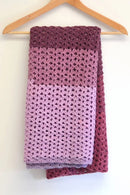 A handmade crocheted scarf with a striped color-blocked pattern in dark plum, dusty rose, and light lavender, hanging from a wooden hanger against a plain white wall.