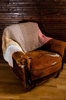 A rustic brown leather armchair draped with a chunky crocheted throw blanket in beige, cream, and soft pink, set against a dark wood paneled wall and light wood floor.