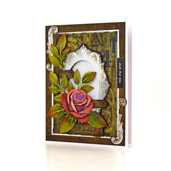 A handmade greeting card featuring a 3D rose with green leaves, an ornate silver frame, and vintage-style elements on a white background.