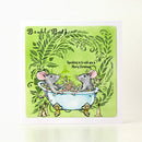 A square Christmas greeting card featuring two gray mice in a light blue bathtub surrounded by green leafy branches, with the text 'Bubble Bath' and 'Squeaking in to wish you a Merry Christmas.' The card is photographed standing upright against a plain white background.