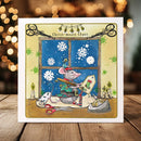 A square greeting card featuring a whimsical illustration of a mouse dressed as Santa Claus sitting by a window with snowflakes outside, candles, scissors, and holiday chaos. The card is displayed standing upright on a wooden surface with a warm, blurred bokeh holiday background.