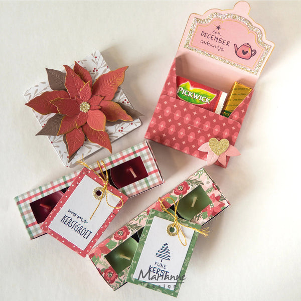 A styled flat-lay of several decorative gift boxes and tags on a light neutral background. The set includes a large poinsettia box, a pink patterned holiday tea box with a gold heart accent, and three smaller patterned boxes with personalized and Christmas-themed paper tags.