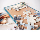 A close-up of a handmade greeting card or art print featuring a jigsaw puzzle design with beach-themed cutout pieces, including a shell piece and a butterfly, on a kraft paper background with a teal border.