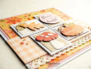 A close-up of a handmade autumn-themed card featuring layered scrapbook papers, die-cut leaf and mushroom shapes, floral accents, and a small circular tag reading 'Here we go.
