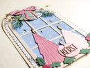 A close-up of a handmade Christmas greeting card featuring a window design with pink snowman figures, green pine branches, gold stars, and a pink circle reading 'KEAST'.