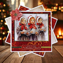 A square Christmas greeting card or art print featuring an illustration of three young girls in winter clothing holding sheets of paper, surrounded by red and gold ornamental borders and a pink decorative flower. The card is stacked on other similar cards on a wooden surface with a blurred holiday background of lights and a warm fireplace.