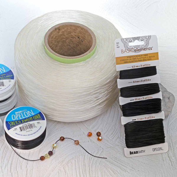 A large spool of white cord or thread sits on a light surface, with several smaller spools and a carded package of black cord labeled BeadSmith Basics Elements OptionL. A few loose beads and a thin cord with beads are placed in front.