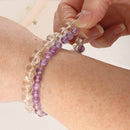 Two beaded stretch bracelets on a wrist: one made of clear round beads and one made of purple round beads, with a hand adjusting the bracelet.