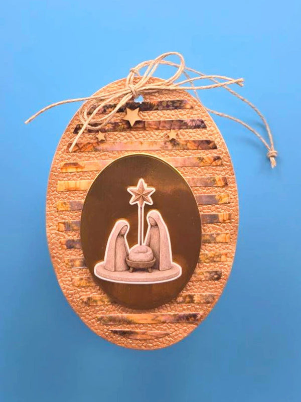An oval wooden hanging ornament with a textured natural wood outer border, a central dark brown oval containing a light wood nativity scene with a star, and a jute hanging string tied at the top, shown against a solid blue background.