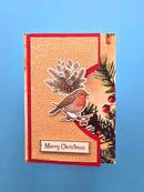 A handmade Christmas greeting card featuring a textured beige background, a red border, a die-cut robin bird, pine branches, red berries, and a 'Merry Christmas' label, displayed against a blue gradient background.