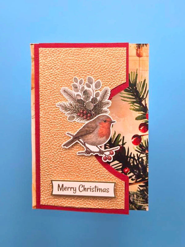 A handmade Christmas greeting card featuring a textured beige background, a red border, a die-cut robin bird, pine branches, red berries, and a 'Merry Christmas' label, displayed against a blue gradient background.