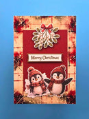 A handmade Christmas greeting card featuring two cute penguins wearing red knit hats and scarves, set against a red textured background with a mistletoe motif and a 'Merry Christmas' banner, displayed on a solid blue background.
