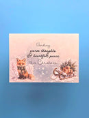 A rectangular Christmas greeting card with a soft pink and blue watercolor background, featuring an illustration of a fox on the left and a sleigh with ornaments on the right. The center contains the handwritten-style text: 'Sending warm thoughts & heartfelt peace this Christmas.