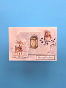 A rectangular Christmas greeting card featuring a soft pink background with illustrated winter motifs, including a deer, a lantern, a bird, and the text 'Merry Christmas', photographed against a solid blue background.