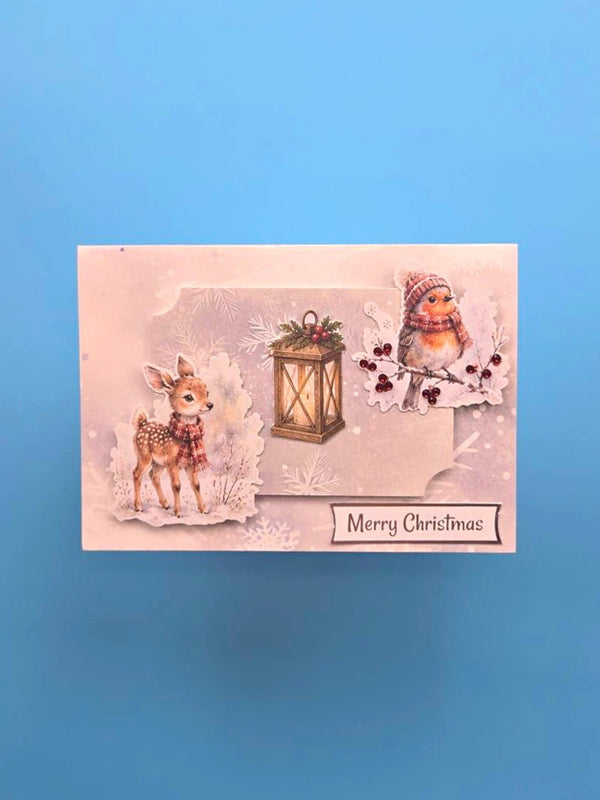A rectangular Christmas greeting card featuring a soft pink background with illustrated winter motifs, including a deer, a lantern, a bird, and the text 'Merry Christmas', photographed against a solid blue background.