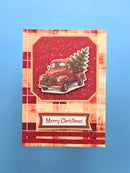 A handmade Christmas greeting card featuring a vintage red pickup truck carrying a Christmas tree, set against a red and beige plaid background with 'Merry Christmas' written on a banner, displayed on a solid blue background.