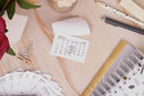 A styled flat-lay of stationery items on a light wood surface, including a small March 2021 calendar page with a pink glittery circle drawn over the 13th, a spiral-bound notebook, paper clips, a pen, and a rose.