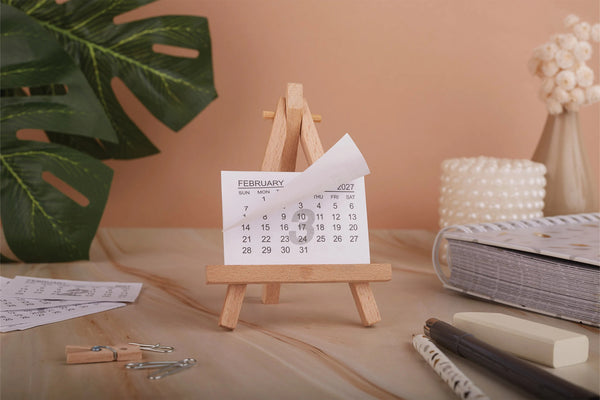 A small wooden calendar stand holding a February calendar page, styled on a tabletop with a notebook, pens, paper clips, a pearl candle, and a green leaf in the background.