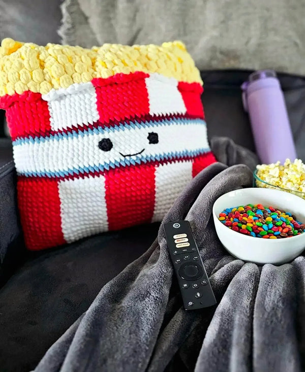 Colorful striped pillow with a face on a couch next to a bowl of popcorn and a remote control.