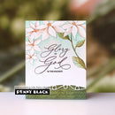 A handmade greeting card with watercolor-style white flowers, green leaves, and the words 'Glory to God' on a white background, standing upright against a blurred green and brown backdrop. A small label at the bottom reads 'Penny Black' and 'papercrafting with Penny Black.