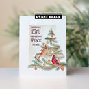 A handmade greeting card featuring a watercolor-style Christmas tree with two cardinals, orange ornaments, and the text 'BORN IN LOVE, BRINGING PEACE TO ALL'. The card is displayed upright on a white surface with a soft-focus background.