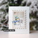 A handmade greeting card featuring a stamped snowman illustration on a postage stamp design, set against a white embossed snowflake background. The card is displayed on a light surface with a blurred winter garden background.