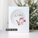 A handmade greeting card with a white textured background, featuring a circular die-cut design with a cute snowman illustration wearing a red Santa hat and holding a small star. The card is displayed on a white surface with a blurred green pine tree in the background.