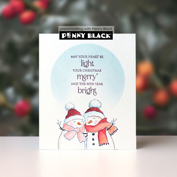 A handmade Christmas greeting card by Penny Black featuring two watercolor snowmen under a light blue circle with the text 'MAY YOUR HEART BE light YOUR CHRISTMAS merry AND THE NEW YEAR bright'. The card is displayed upright on a white surface with a blurred festive background of pine branches and red berries.