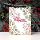 A Penny Black paper crafting stamp card featuring a festive Christmas design with eucalyptus leaves, red berries, and the text 'Rejoice THIS HOLY SEASON.' The card is displayed upright against a blurred background of pine branches and red berries.