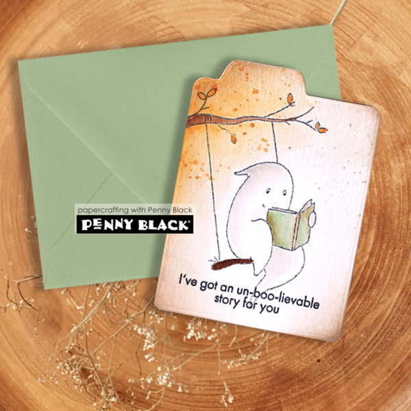 A handmade greeting card with a cute ghost illustration reading a book, placed on top of a sage green envelope on a wooden surface. The card has a visible 'Penny Black' stamp brand label and the text 'I've got an un-boo-liable story for you'.