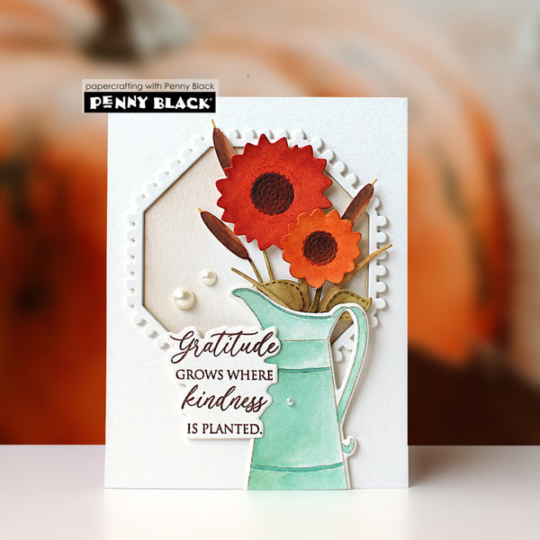 A handmade greeting card by Penny Black featuring a white background with a die-cut white frame, a mint green pitcher, orange and red flowers, and the text 'Gratitude GROWS WHERE kindness IS PLANTED.' The card is photographed standing upright against a softly blurred autumn-themed background.