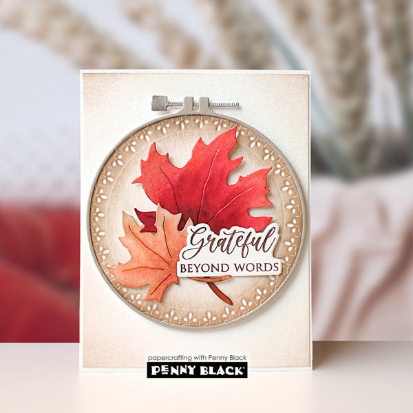 A handmade greeting card featuring a circular layered design with red and orange maple leaves, a beige scalloped border, and the words 'Grateful BEYOND WORDS.' The card is shown upright against a softly blurred warm-toned background, with the Penny Black branding visible at the bottom.