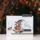 A handmade Halloween greeting card by Penny Black featuring a cute cartoon witch in a black hat holding a pumpkin, with layered white 'BOO' letters, a ghost, and a bat against a dark blurred background with warm lights.