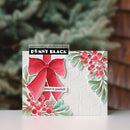 A handmade greeting card featuring a large red bow, green leaves, and red berries on a white embossed background, with a black banner reading 'PENNY BLACK' and a small tag that says 'peace & goodwill'. The card is photographed standing upright on a white surface with a blurred evergreen and brick background.