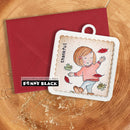 A handmade autumn-themed greeting card featuring a cartoon girl with falling leaves, shown with a red envelope on a wood slice background. The card has a white scalloped border and a paperclip hole at the top.