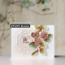 A handmade Christmas card featuring pinecones, holly leaves, and red berries layered over a white geometric frame with the text 'JOY IN CHRIST'. The card is displayed on a light surface with a blurred background, and the brand 'Penny Black' is visible on the packaging label.