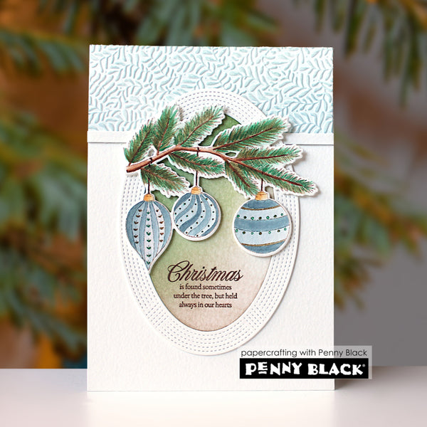 A handmade Christmas greeting card featuring three patterned blue and white ornaments, pine branches, and a quote, displayed against a blurred green background with a Penny Black brand label at the bottom.
