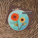A handmade circular embroidery-style card or art piece featuring two layered flower motifs in orange and yellow on a turquoise background, with the word 'Gratitude' and pearl embellishments. It is displayed in a wooden hoop over a brown envelope on a woven wicker surface, with a Penny Black stamp brand sticker visible.