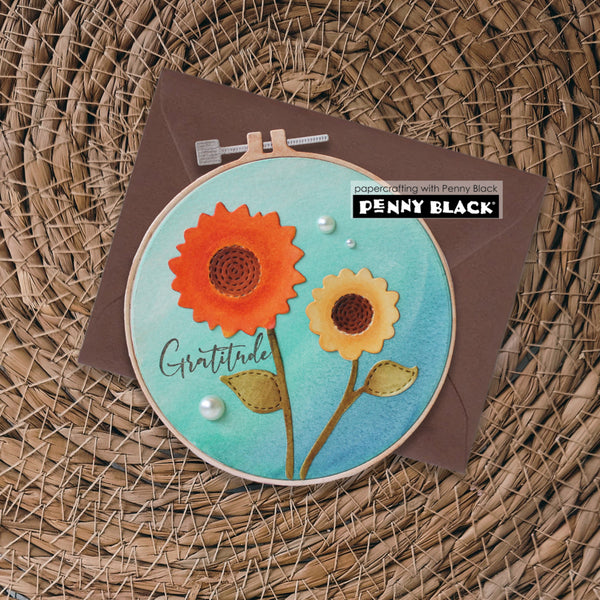 A handmade circular embroidery-style card or art piece featuring two layered flower motifs in orange and yellow on a turquoise background, with the word 'Gratitude' and pearl embellishments. It is displayed in a wooden hoop over a brown envelope on a woven wicker surface, with a Penny Black stamp brand sticker visible.