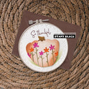 A handmade greeting card featuring a watercolor pumpkin with colorful flowers, styled in an embroidery hoop on top of a brown envelope, set against a woven wicker background. The card includes the text 'So thankful' and a 'papercrafting with Penny Black PENNY BLACK' logo.