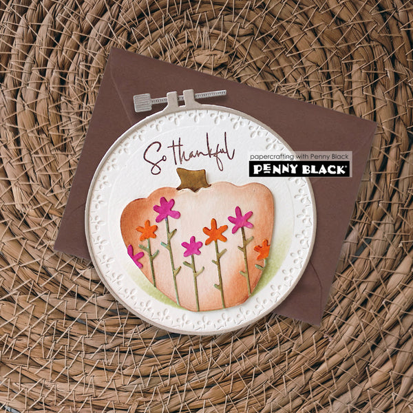A handmade greeting card featuring a watercolor pumpkin with colorful flowers, styled in an embroidery hoop on top of a brown envelope, set against a woven wicker background. The card includes the text 'So thankful' and a 'papercrafting with Penny Black PENNY BLACK' logo.