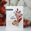 A handmade layered paper card featuring a stylized tree with red and brown leaves, stacked books, a small pumpkin, and the text 'have a spook-tacular Halloween.' The card is photographed upright on a white surface with a softly blurred autumn-themed background.