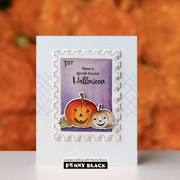 A handmade white greeting card featuring a die-cut stamp design with two carved pumpkins, a purple background, and the text 'have a spooky-tacular Halloween.' The card is photographed upright against a blurred orange autumn background, with a Penny Black brand label visible at the bottom.