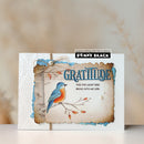 A handmade greeting card featuring a bluebird on a branch with the word 'GRATITUDE' and the quote 'FOR THE LIGHT YOU BRING INTO MY LIFE.' The card has layered rustic paper textures, blue painted edges, and a small black Penny Black stamp label near the top.