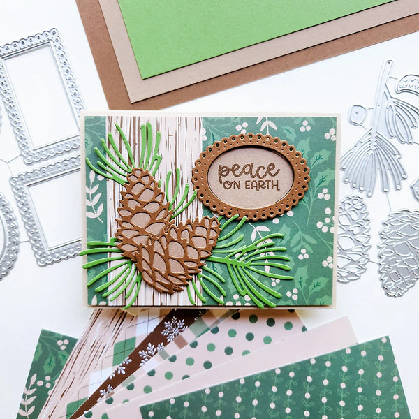 A handmade layered card featuring pinecones and green pine needles on a wood-grain and green patterned background, with a scalloped oval label reading 'peace ON EARTH.' Silver die-cut embellishments and patterned paper sheets are arranged around it on a white surface.