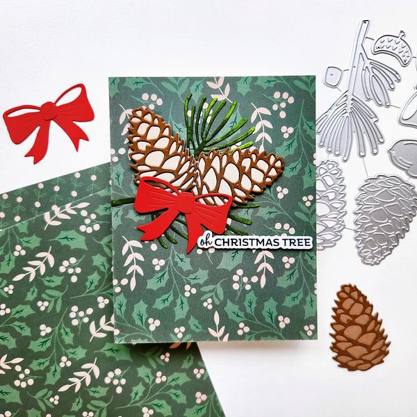 A handmade Christmas card with a green holly and berry pattern, featuring a layered brown pinecone and red bow design. The card is displayed on a white background alongside matching patterned paper, silver metal die-cutting tools, and a red paper bow.