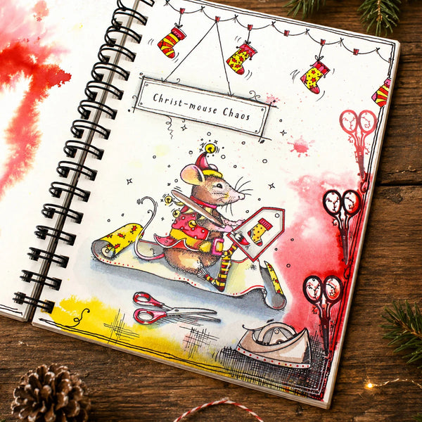 A spiral-bound notebook or journal page featuring a whimsical Christmas mouse illustration riding a sleigh, with stockings, scissors, and the text 'Christ-mouse Chaos' on a rustic wooden surface.