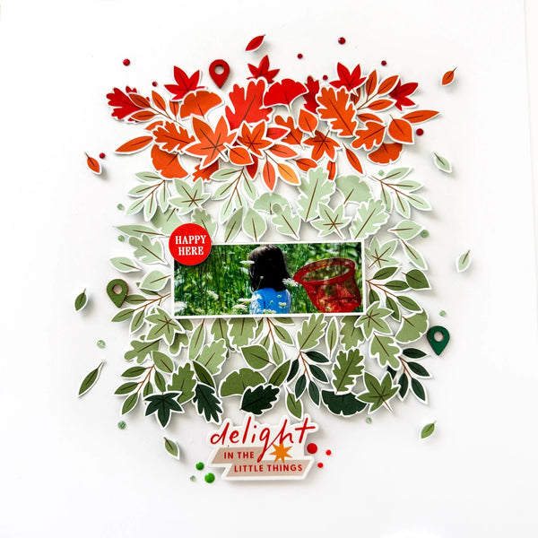 A layered scrapbook-style paper collage on a white background with red and orange autumn leaves at the top, green leaves surrounding a central photo insert, and the phrase 'delight in the little things' near the bottom.