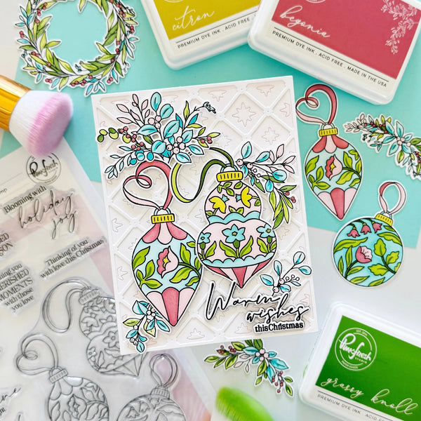 A handcrafted Christmas card featuring two colorful ornament illustrations with floral details and the text 'Warmest wishes this Christmas,' styled on a mint background with ink pads, stamps, and brushes.