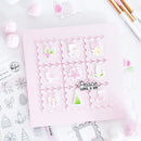A handmade paper advent calendar card in light pink, featuring nine postage-stamp-style windows with winter-themed illustrations like a deer, bell, bird, flower, candle, gift, Christmas tree, stocking, and the words 'Peace, Love & Joy'. The card is styled on a white surface with craft supplies, pom-poms, and clear stamps.